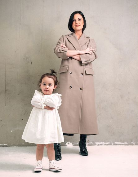 Rochelle standing in a light brown full length coat with black boots on, with her daughter in the foreground in a white dress and white shoes, both with their arms cross