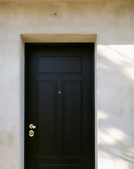 A white stone wall with a large black wooden door, with bronze handle on the left side and keyhole below